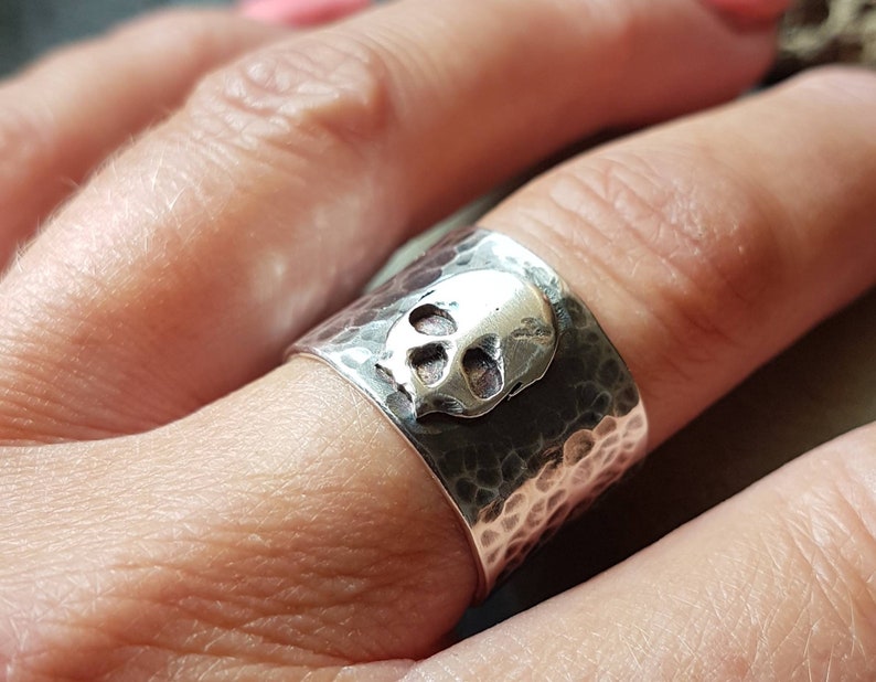 May include: A silver ring with a hammered finish and a skull design. The ring is worn on a finger.