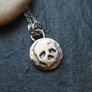 May include: A silver necklace with a small, round pendant featuring a skull design. The pendant has a textured surface and the skull is raised.