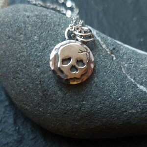 May include: A silver necklace with a small, hammered skull pendant. The pendant is round and has a detailed skull design.