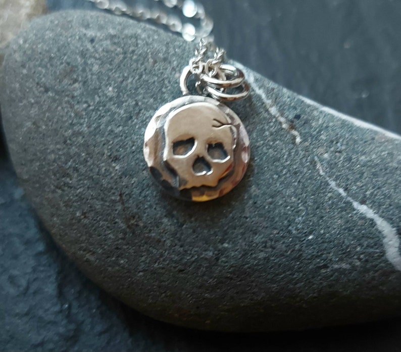 May include: A silver necklace with a small, hammered skull pendant. The pendant is round and has a detailed skull design.