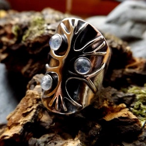 May include: A silver ring with a tree design and three moonstone gems. The ring is set on a piece of wood.