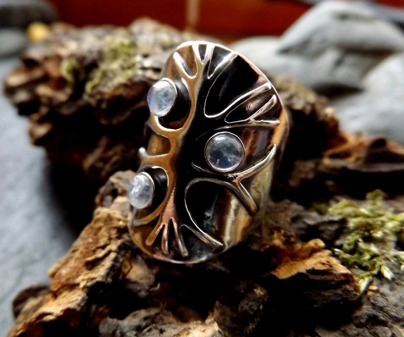 May include: A silver ring with a tree design and three moonstone gems. The ring is set on a piece of wood.