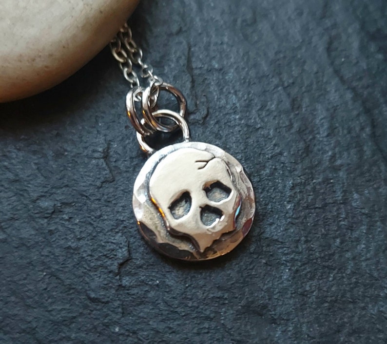 May include: A silver necklace with a small, round pendant featuring a skull design. The pendant has a textured surface and the skull is raised.