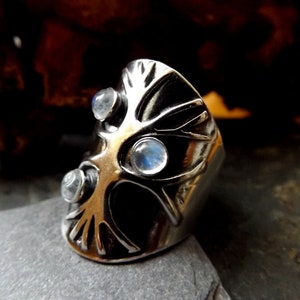 May include: A silver ring with a tree design and three moonstone gems. The ring is set on a dark gray surface.