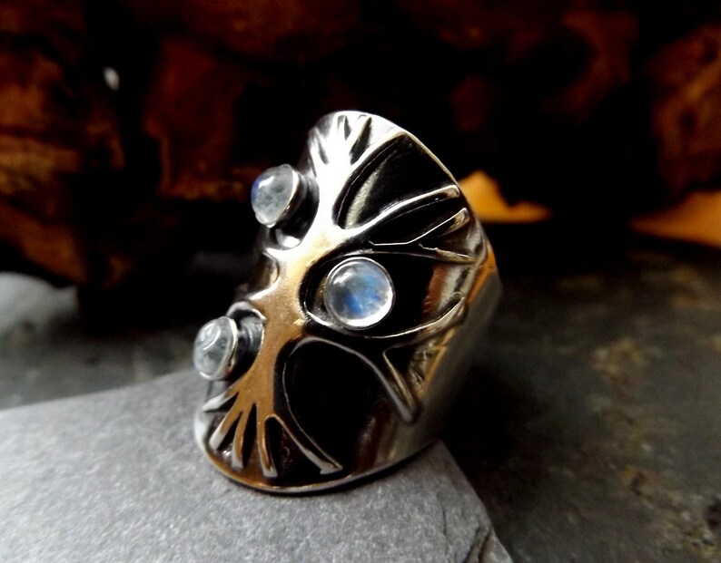 May include: A silver ring with a tree design and three moonstone gems. The ring is set on a dark gray surface.