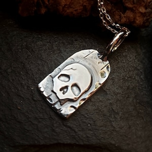 May include: Silver pendant necklace with a skull design. The pendant is rectangular and has a textured surface. The skull is raised and has a detailed design.