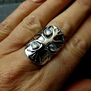 May include: A silver ring with a tree design and three moonstone gems. The ring is worn on a finger.