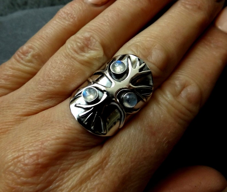 May include: A silver ring with a tree design and three moonstone gems. The ring is worn on a finger.