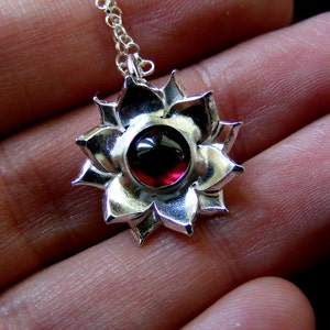 May include: A silver pendant necklace with a red gemstone in the center, shaped like a flower with multiple petals.