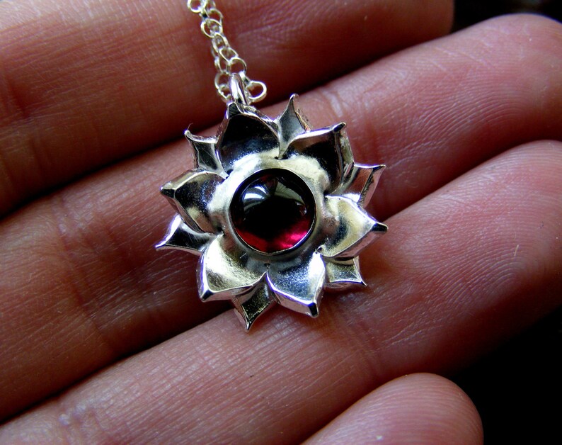 May include: A silver pendant necklace with a red gemstone in the center, shaped like a flower with multiple petals.