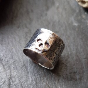 May include: A silver skull ring with a hammered texture. The skull is raised and detailed.