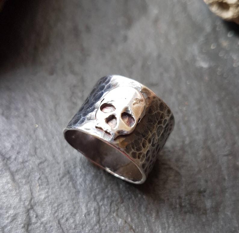 May include: A silver skull ring with a hammered texture. The skull is raised and detailed.