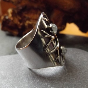 May include: A silver ring with a unique design featuring a branch-like pattern and two clear gemstones. The ring is wide and has a rounded band.
