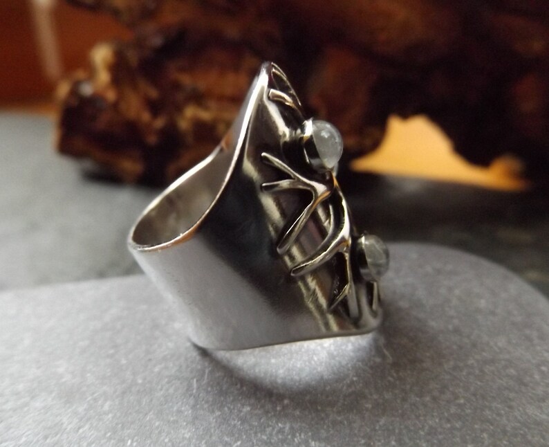 May include: A silver ring with a unique design featuring a branch-like pattern and two clear gemstones. The ring is wide and has a rounded band.