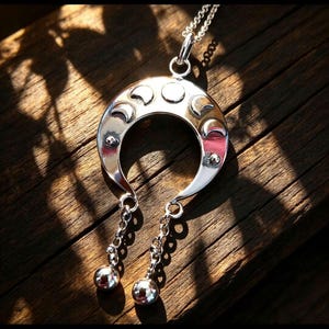 May include: A silver-toned crescent moon pendant necklace featuring a series of moon phase engravings along the curved top edge. Two short silver chains hang from the bottom of the crescent, each ending in a small, polished silver sphere.