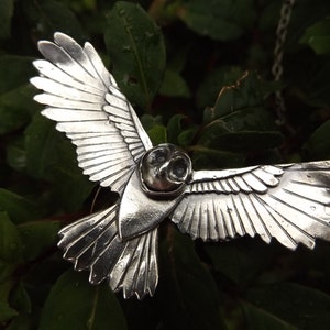 May include: A silver owl pendant with detailed wings, a hollowed-out head, and black eyes. The pendant is hanging from a silver chain.