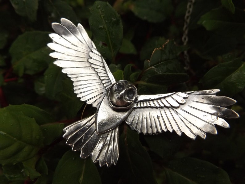 May include: A silver owl pendant with detailed wings, a hollowed-out head, and black eyes. The pendant is hanging from a silver chain.