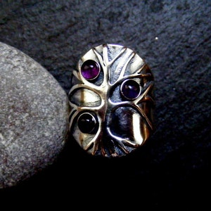 May include: A silver ring with a tree design and three purple gemstones. The ring is on a gray background.