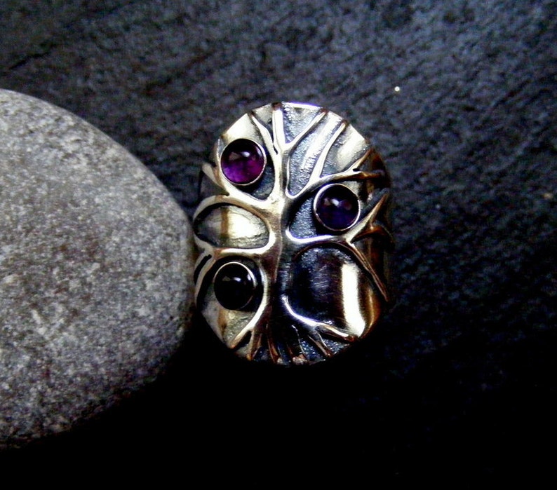 May include: A silver ring with a tree design and three purple gemstones. The ring is on a gray background.