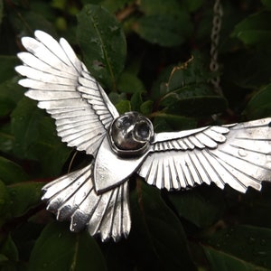 May include: A silver owl pendant with detailed wings spread wide, hanging from a silver chain. The owl's eyes are black and the pendant is set against a green leafy background.