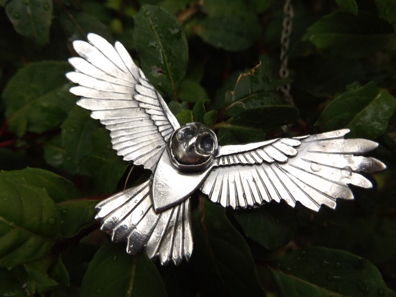May include: A silver owl pendant with detailed wings spread wide, hanging from a silver chain. The owl's eyes are black and the pendant is set against a green leafy background.