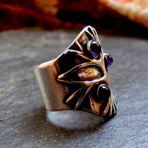 May include: A silver ring with a geometric design and three purple gemstones. The ring is set on a gray background.