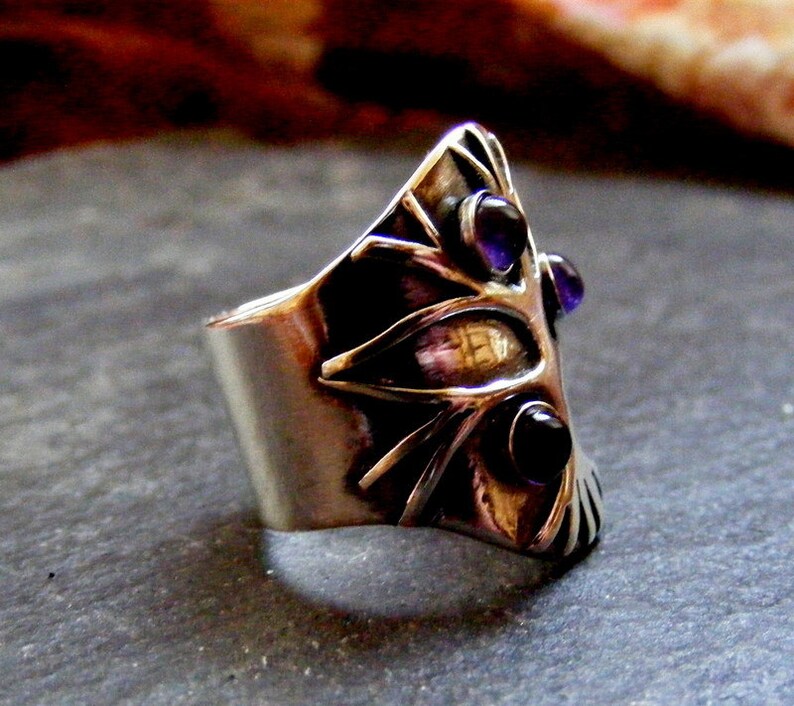 May include: A silver ring with a geometric design and three purple gemstones. The ring is set on a gray background.