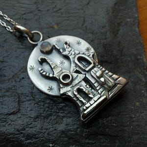 May include: A silver pendant necklace featuring a circular design with two tall, pointed structures resembling towers. The background of the pendant is etched with small star shapes and a single round, smooth stone set near the top edge.
