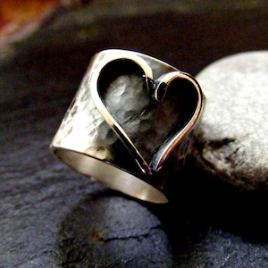 May include: A silver ring with a hammered texture and a heart-shaped cutout. The heart is outlined with a gold-colored metal. The ring is set against a dark background.