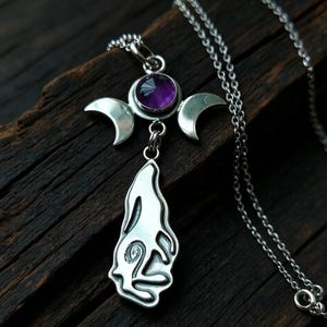 May include: A silver-toned pendant necklace featuring a central circular purple gemstone flanked by two crescent moon shapes. Below the moons hangs a stylized silver-toned figure of a howling wolf with a spiral detail on its body.