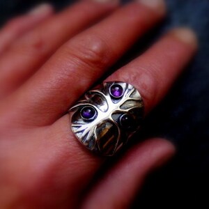 May include: A silver ring with a tree design and three purple gemstones. The ring is worn on a person's finger.