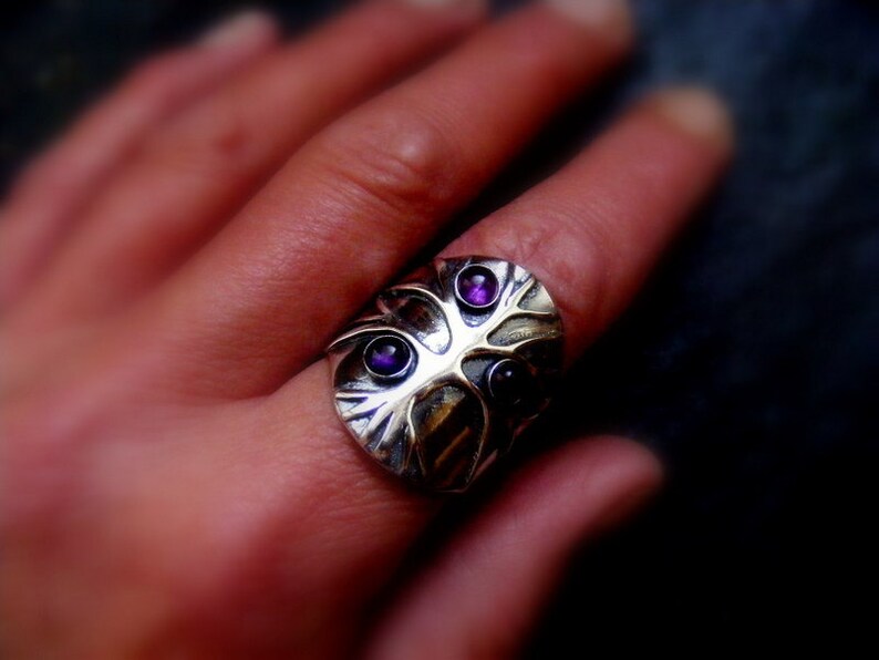 May include: A silver ring with a tree design and three purple gemstones. The ring is worn on a person's finger.
