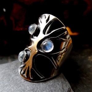 May include: A silver ring with a tree design and three moonstone gems. The ring is wide and has a textured surface.