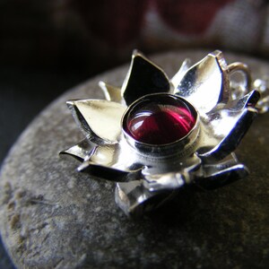 May include: A silver pendant in the shape of a flower with a red gemstone in the center.