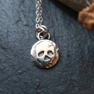 May include: A silver necklace with a small, round pendant featuring a skull design. The pendant has a textured surface and the skull is raised and detailed.