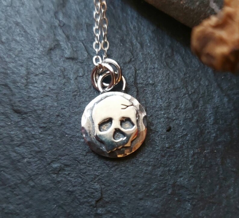 May include: A silver necklace with a small, round pendant featuring a skull design. The pendant has a textured surface and the skull is raised and detailed.