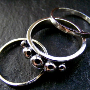 Puede incluir: Tres anillos de plata apilados uno encima del otro. El anillo inferior es liso, el anillo del medio tiene una fila de cinco protuberancias redondas y el anillo superior es liso.
