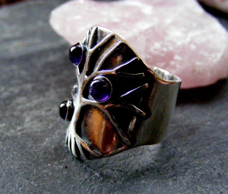 May include: A silver ring with a tree design and three amethyst stones. The ring is set on a gray background.