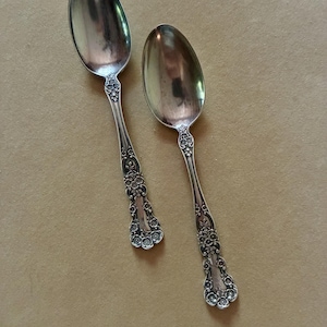 May include: Two silver-toned metal spoons with ornate floral detailing on the handles. The spoons feature a polished oval bowl and a decorative stem with embossed flower patterns that extend from the neck down to the rounded handle end.