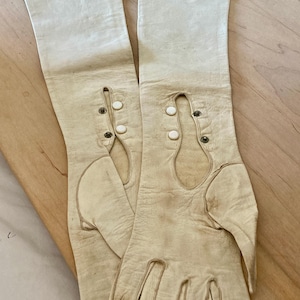 May include: A pair of off-white, vintage-style gloves. The gloves are long, extending past the wrist, and feature decorative button details. The leather appears aged, with some discoloration, suggesting they are antique or vintage.
