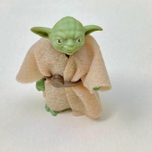 May include: A green Yoda figurine with large ears, wearing a tan robe and a brown belt. The Star Wars character has a wrinkled face and is standing on a white surface. The figurine is a collectible item.