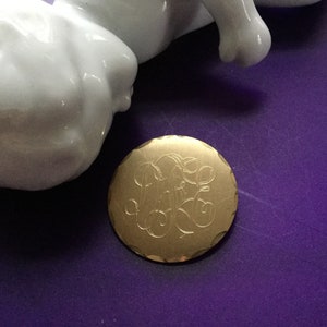 May include: A gold-toned round brooch with a monogrammed design. The monogram is engraved into the metal and features the letters "M.E.G."