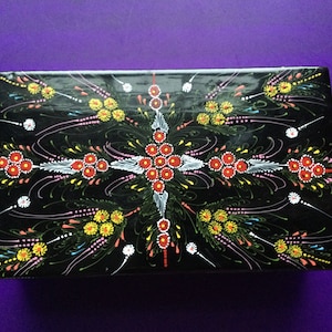 May include: A black wooden box with a hand-painted design. The design features a symmetrical pattern of red, yellow, and white flowers on a black background.