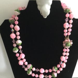 May include: A double strand pink and green glass bead necklace with a floral design. The necklace is on a black display stand.
