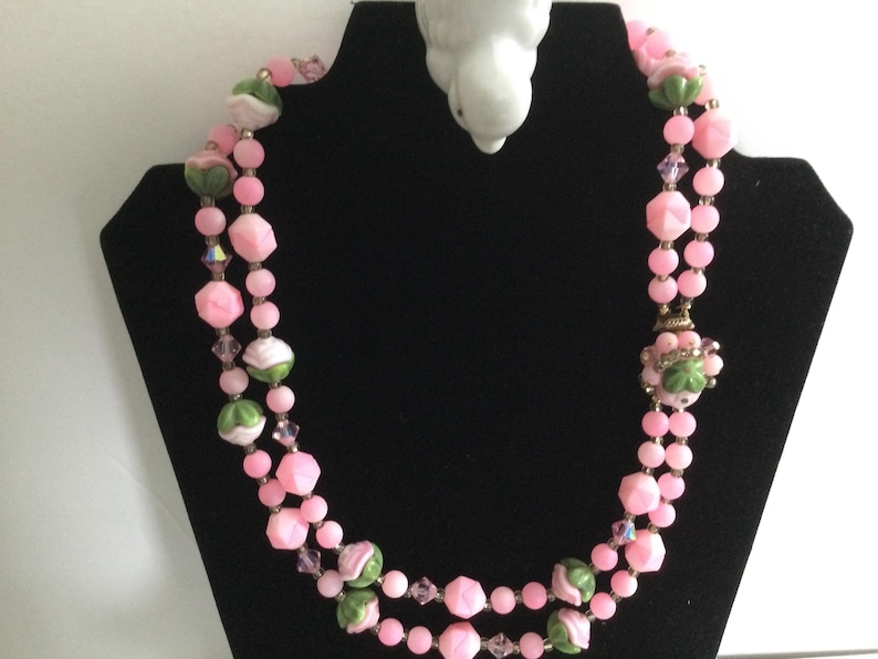 May include: A double strand pink and green glass bead necklace with a floral design. The necklace is on a black display stand.