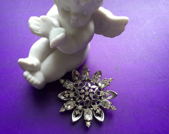 Könnte beinhalten: Eine silberne Brosche in Form einer Schneeflocke mit klaren Strasssteinen auf einem violetten Hintergrund. Die Brosche liegt neben einer weißen Porzellanfigur eines Engels.