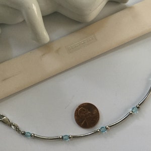 May include: A silver bracelet with a delicate chain and light blue gemstone accents. The bracelet is displayed next to a penny for size reference. The brand name "Swarovski" is visible on a white tag.