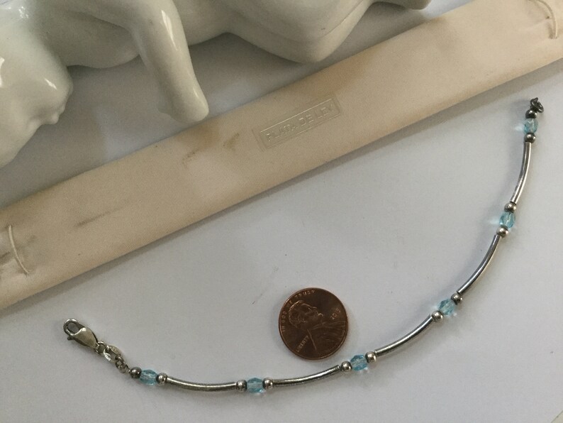 May include: A silver bracelet with a delicate chain and light blue gemstone accents. The bracelet is displayed next to a penny for size reference. The brand name "Swarovski" is visible on a white tag.