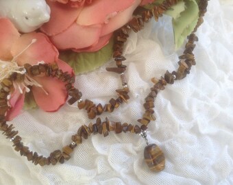 May include: A brown tiger's eye stone necklace with a pendant. The necklace is made of small, irregular shaped stones and is strung on a silver chain. The pendant is a larger, oval-shaped tiger's eye stone.