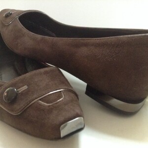 May include: A pair of brown suede flats with a decorative button on the toe. The shoes have a silver trim around the toe and heel.